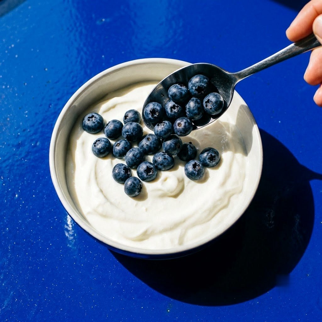 Blueberries being spooned into a bowl of yoghurt on a blue worktop